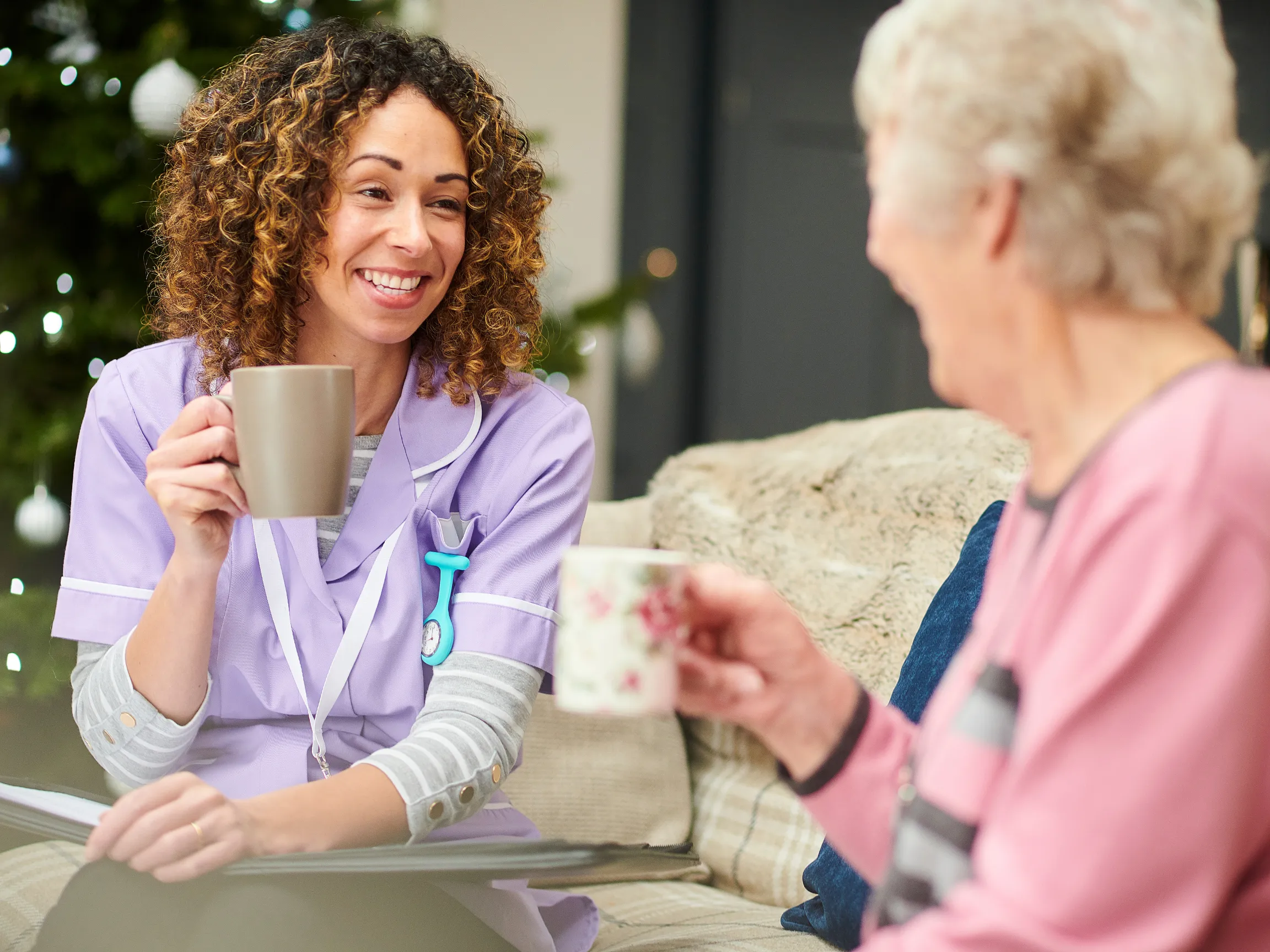 elderly woman and caregiver sipping coffee in front of a christmas tree during in-home care during the holidays